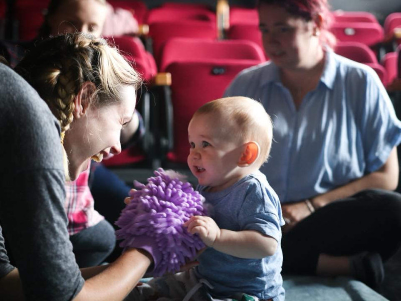 a smiling performer puppets a woolly purple hand towards a baby who touches the purple puppet and smiles at the performer