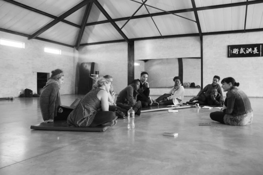 a black and white image of a rehearsal room in Cape Town. 9 young performers sit on mats having a conversation with Anna