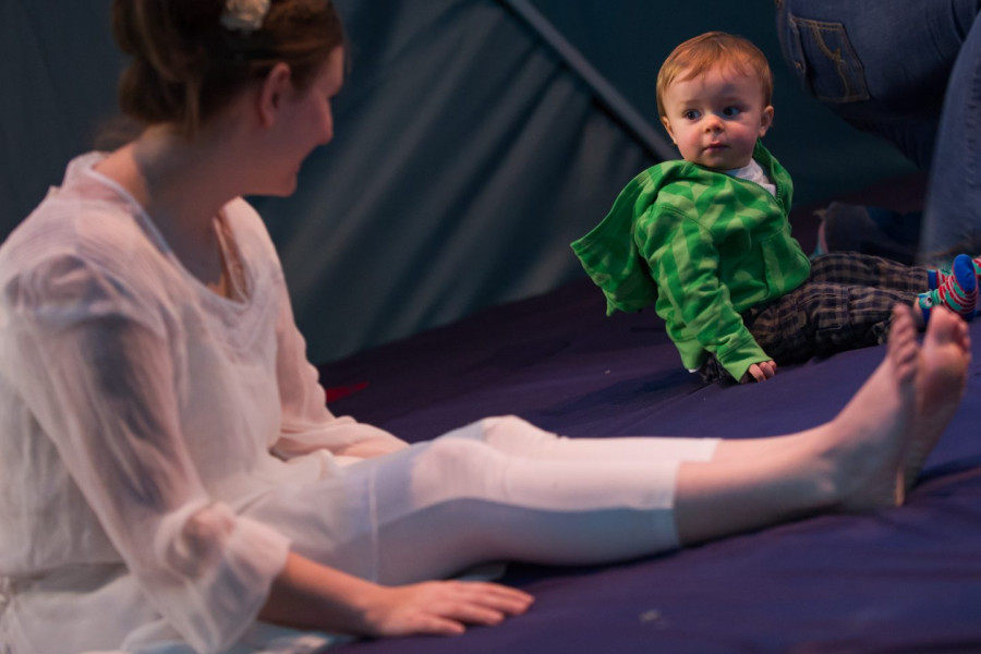Babble - a performer in white sits on a blue floor copying the position of a baby who regards her