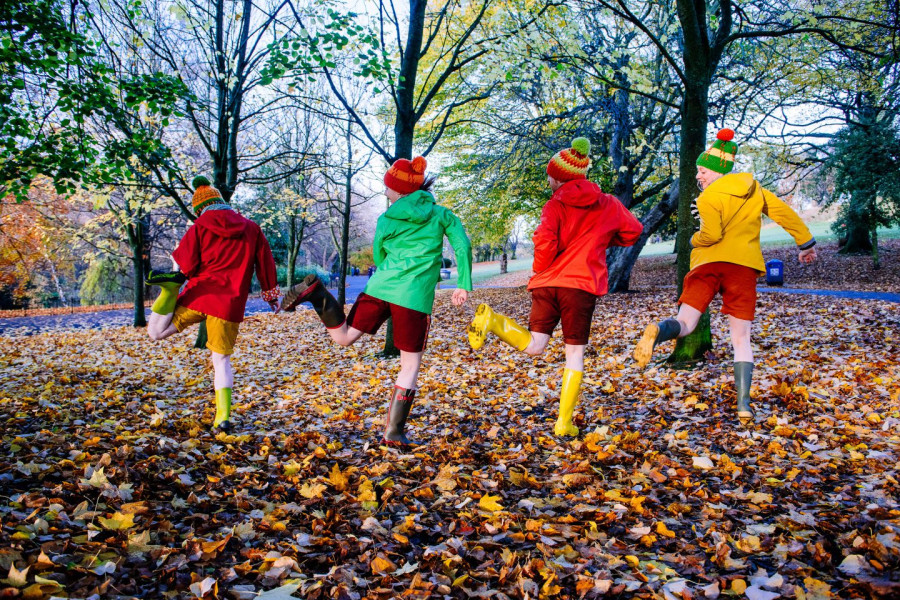 4 Go Wild In Wellies - four performers in bobble hats, raincoats, shorts and wellies in an autumnal park setting with lots of leaves on the ground. their backs to us and they are kicking one leg up each to look at their wellies