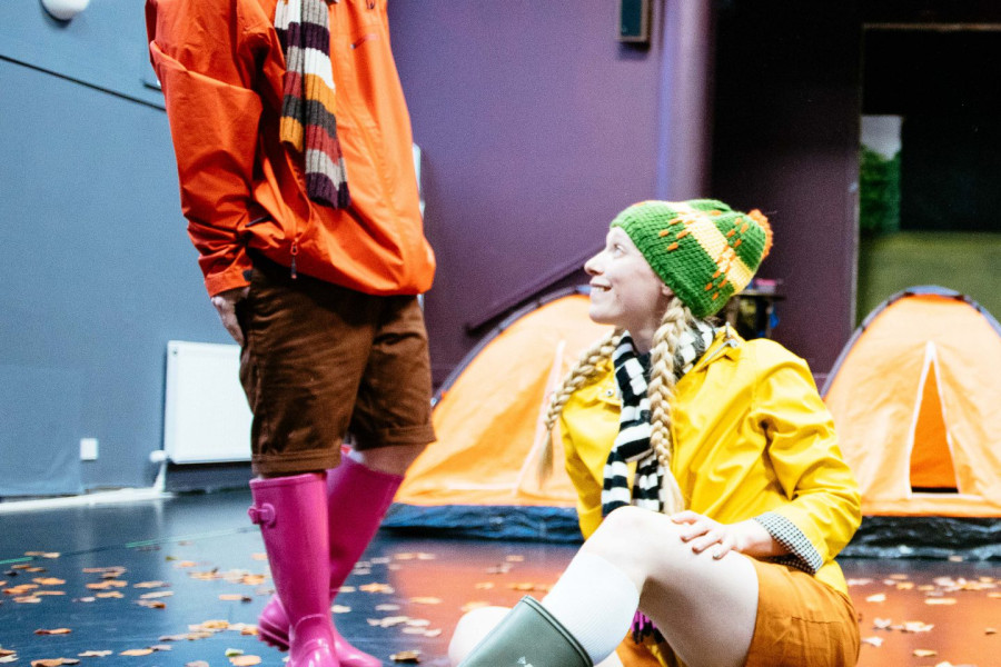 4 Go Wild In Wellies - a performer in an orange bobble hat, stripy scarf, raincoat and wellies stands smiling down at another performer who sits on the floor. she wears a green bobble hat, yellow raincoat, shorts and wellies. there are two orange tents in the background