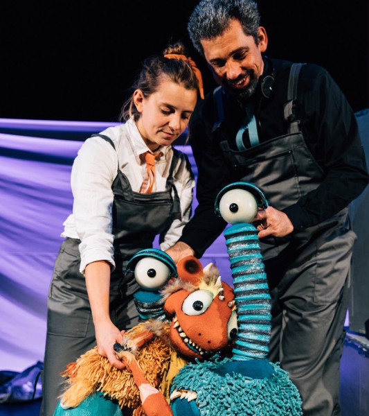 Little Monster - two performers puppeting two monsters - one orange monster with spikey teeth, one round blue monster with eyes on stalks. the monsters embrace