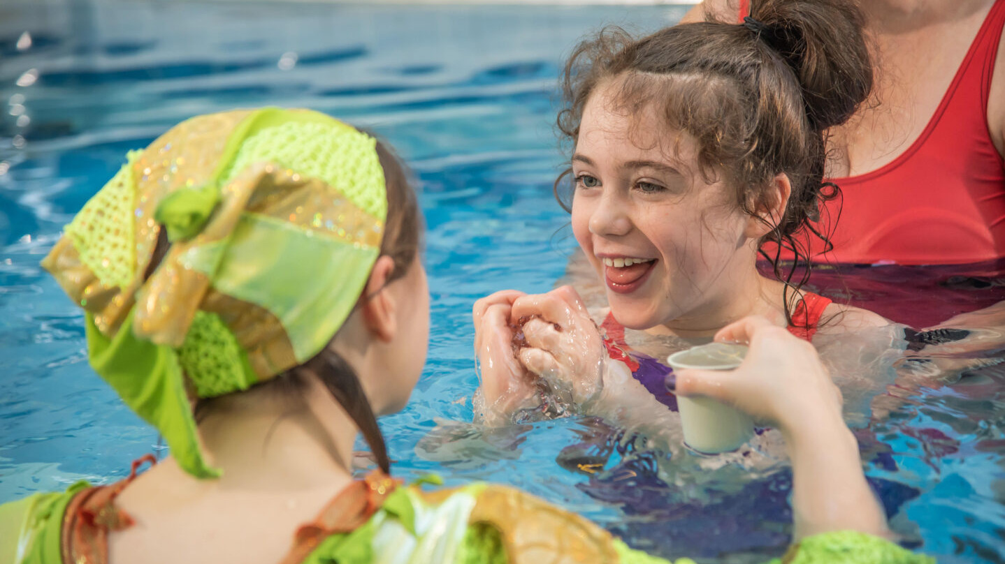 Sing Me To The Sea - a young female audience member smiles at the performer in green who has a small pot of water that she is trickling into the pool. the child is supported by an adult. they are all in a swimming pool