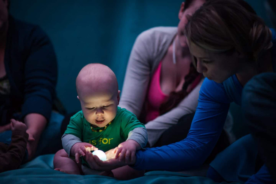 Tiny - a performer holds a tiny light in her palm. a baby looks intently at it and touches her hand. in the background a mother watches