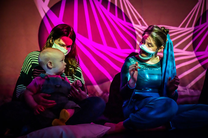 white lines projected on a purple background. in front sits a performer in blue who moves her hands and captivates the baby next to her