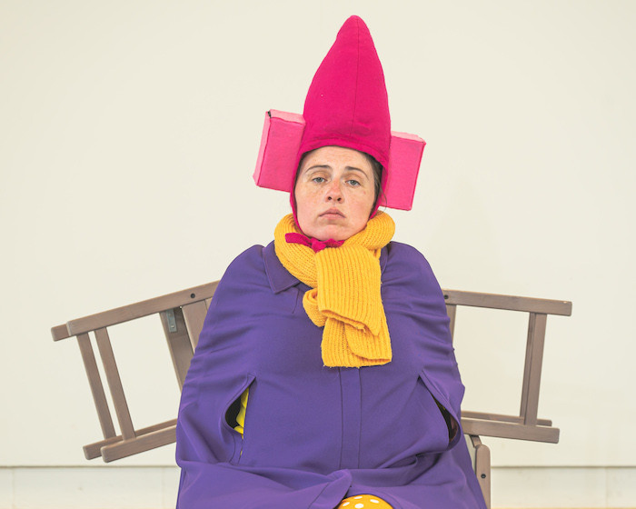 A character in a purple cape and a pink pointed hat sits looking bored. two chairs stick out from her shoulders