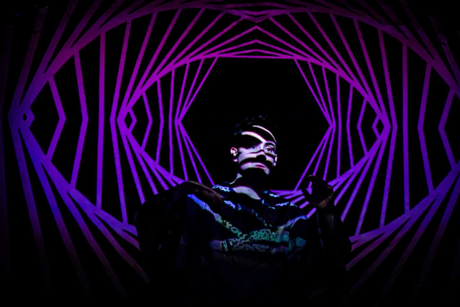 a performer stands in the darkness lit by geometric images projected onto him