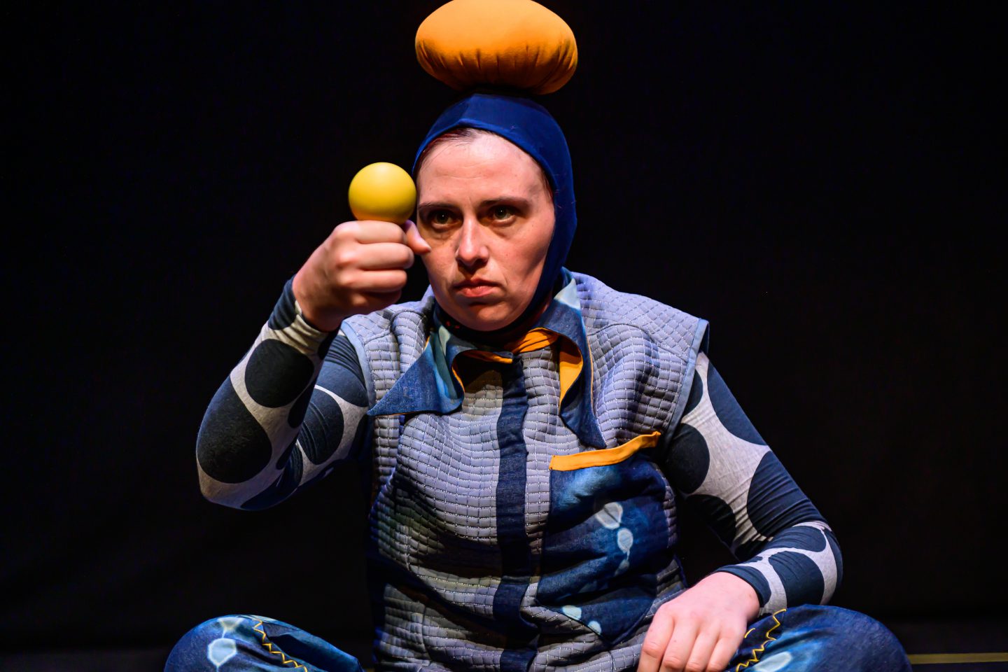 Bigkidlittlekid - a performer in blue with a hat with a giant bobble. she balances a yellow ball on her fist and looks intently at it
