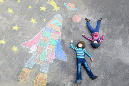 two children in stripy shirts and jeans lie on a playground where there are drawings in chalk of a rocket, stars and planets