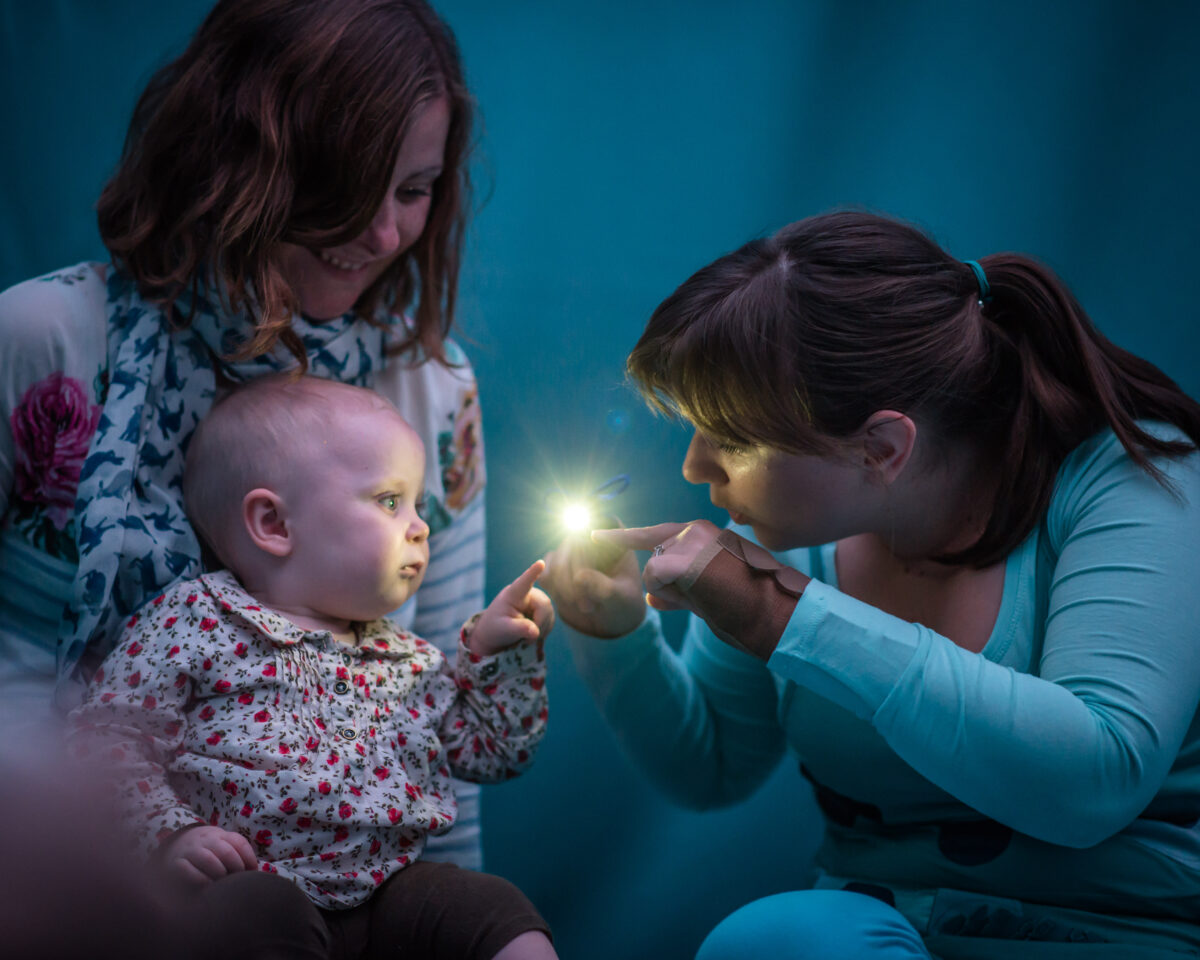 TiNY - a performer in blue holds out a tiny light that a baby reaches to touch. the mother smiles in the background