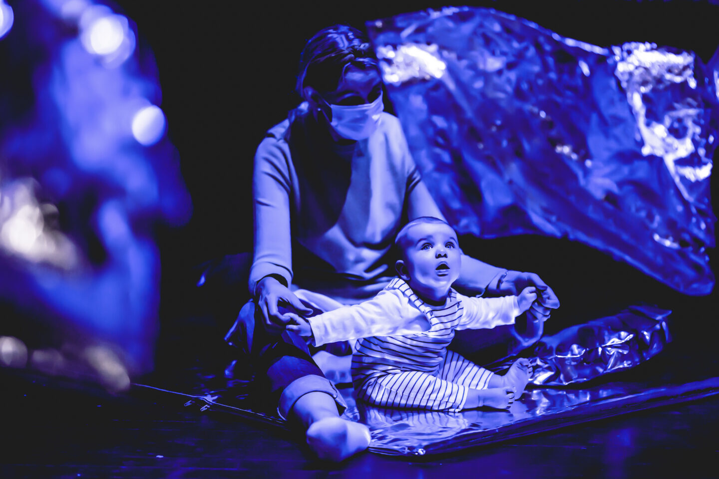 a baby sits on a silver cushion staring at silver foil dancing in front of it. everything is saturated in blue