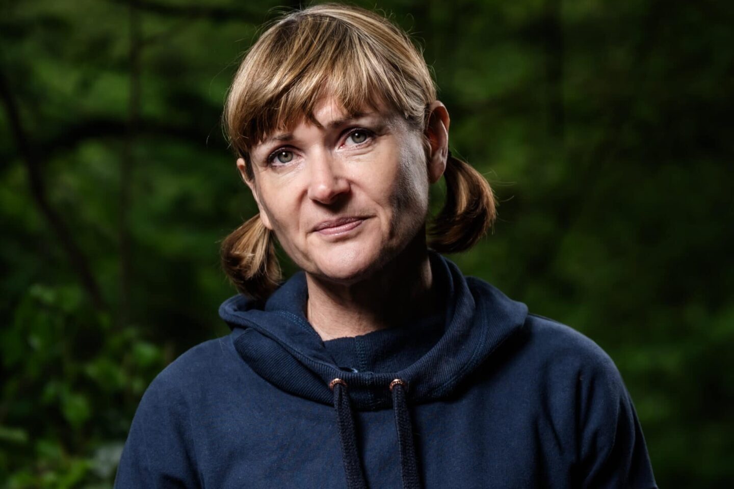 a white woman with short bunches and a navy hoodie looking at the camera against a leafy background
