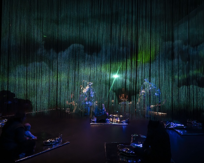through the string drape of the set of this show, we dimly see the outline of performers dancing wearing silvery capes. traces of images of clouds are on the drapes and we can just see the babies in the audience and their adults sitting on silver mats spread out over the stage