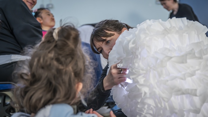 a performer holds a lit up giant ball of crinkled fabric. we see the back of a young woman's head who watches with interest